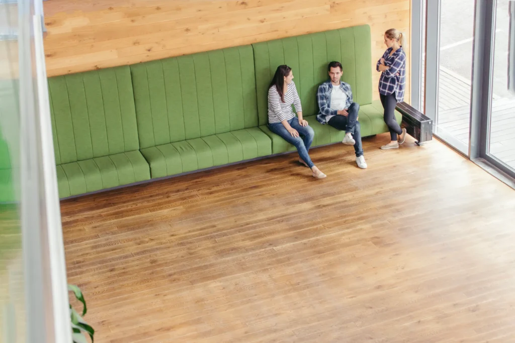 Large modern room with wood flooring and green upholstered bench seating where three people are talking