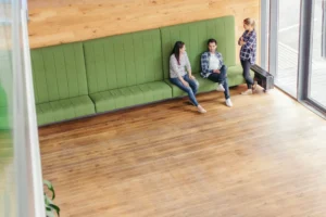 Large modern room with wood flooring and green upholstered bench seating where three people are talking