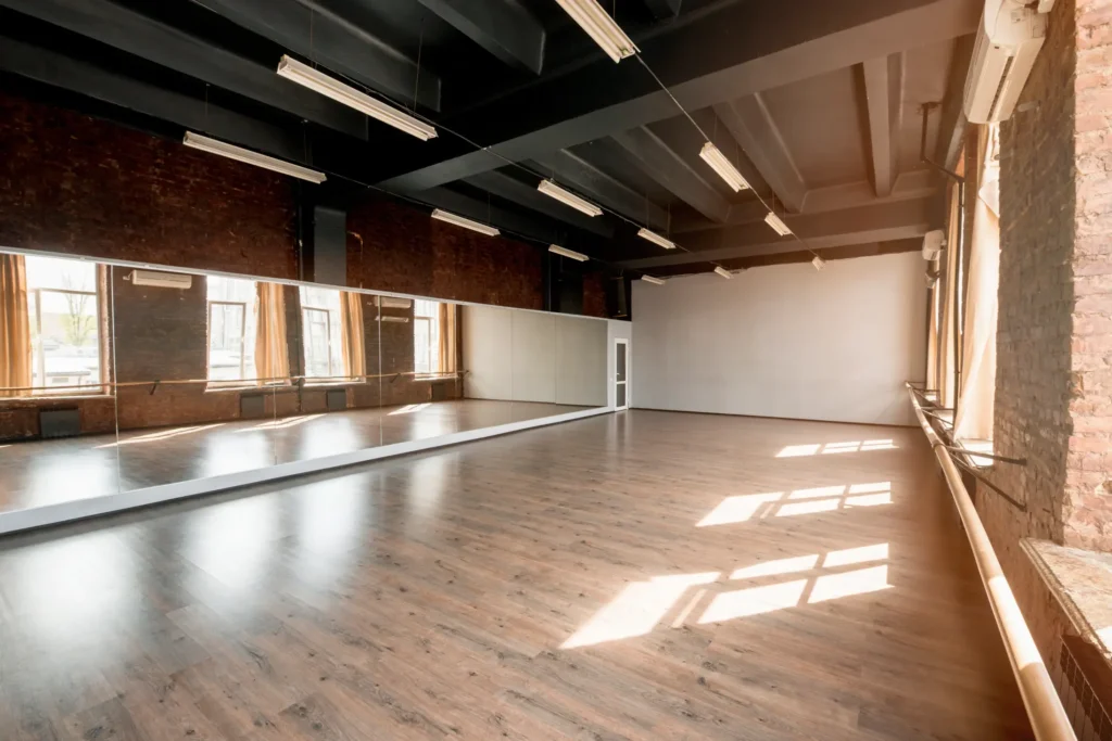 Spacious dance or fitness studio with floor-to-ceiling mirrors, wood flooring, and industrial ceiling lights.