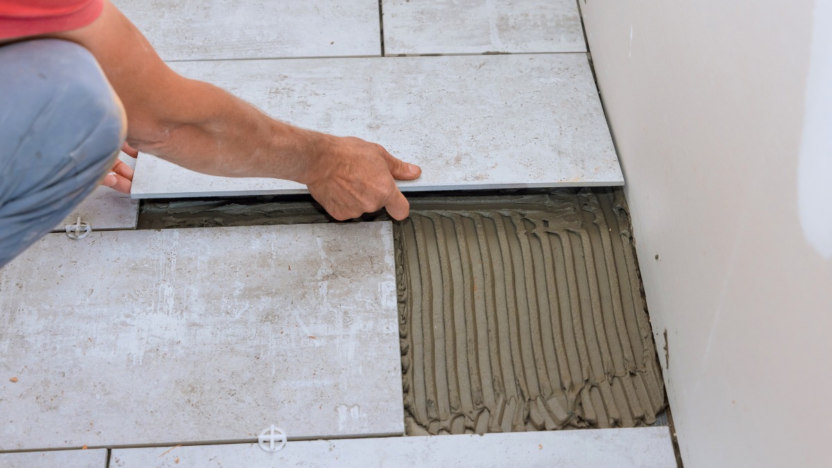 A tiler placing a light grey ceramic floor tile onto combed mortar bedding